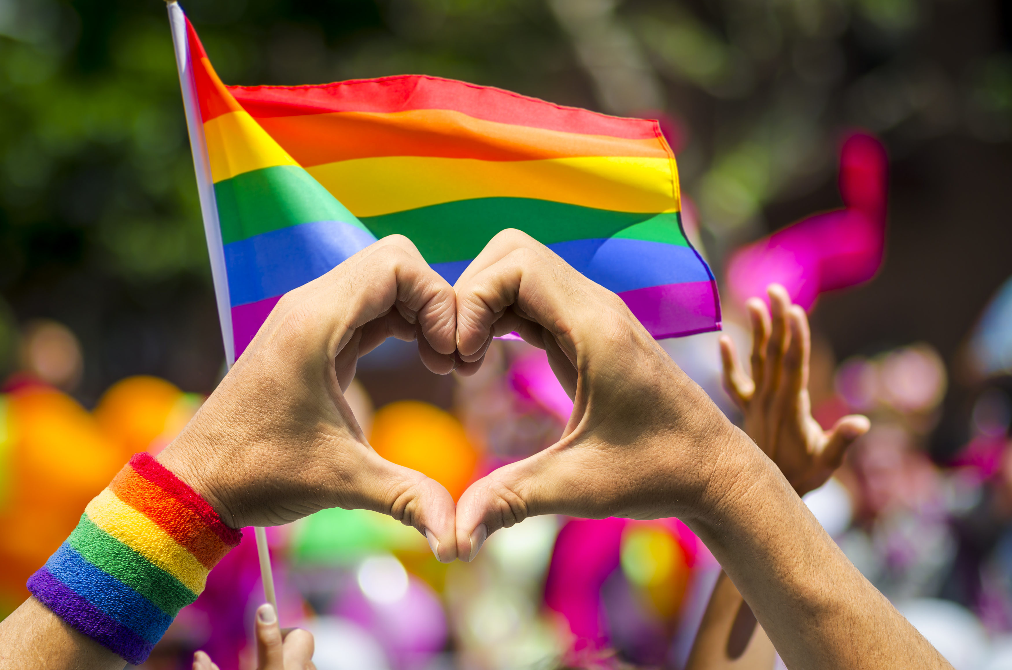 Supporting hands make heart sign and wave in front of a rainbow flag flying on the sidelines of a summer gay pride parade