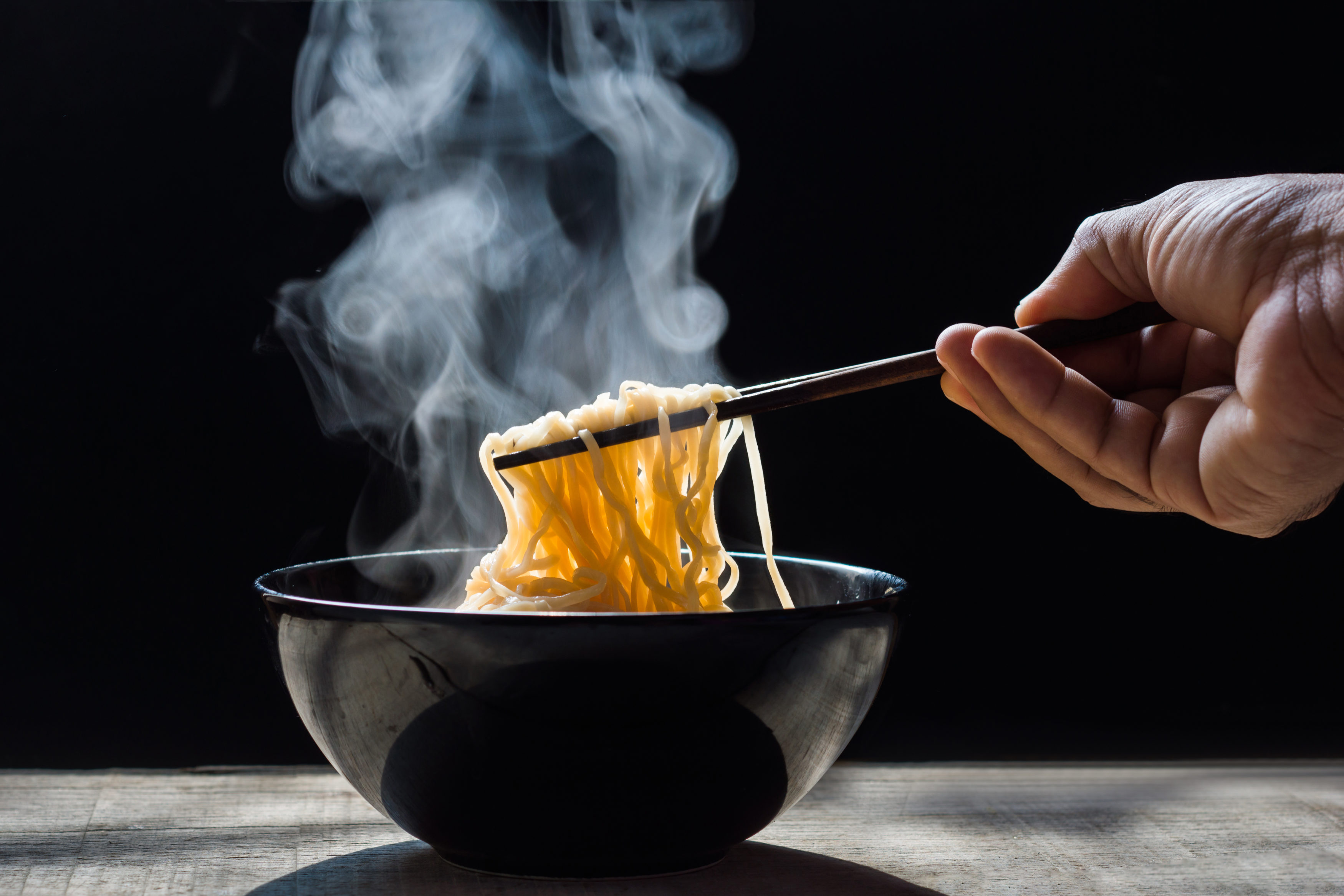 Hand uses chopsticks to pickup tasty noodles with steam and smoke in bowl on wooden background, selective focus.  Top view, Asian meal on a table, junk food concept