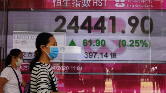 People wearing face masks walk past a bank electronic board showing the Hong Kong share index at Hong Kong Stock Exchange Monday, April 20, 2020. Shares were mixed in Asia on Monday, while oil prices have fallen back. (AP Photo/Vincent Yu)