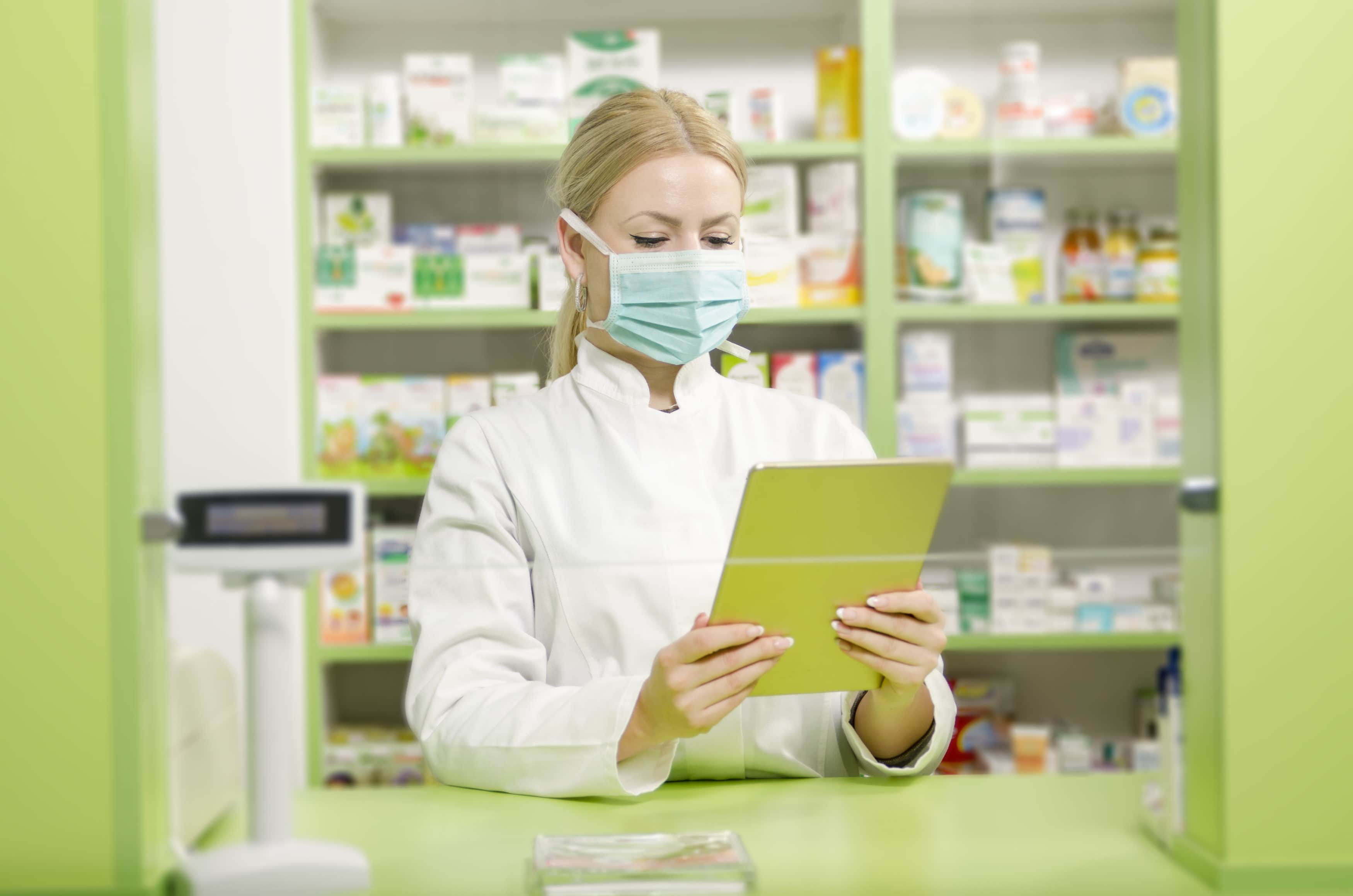 Female pharmacist with surgical mask behind cash register holding tablet in her hands
