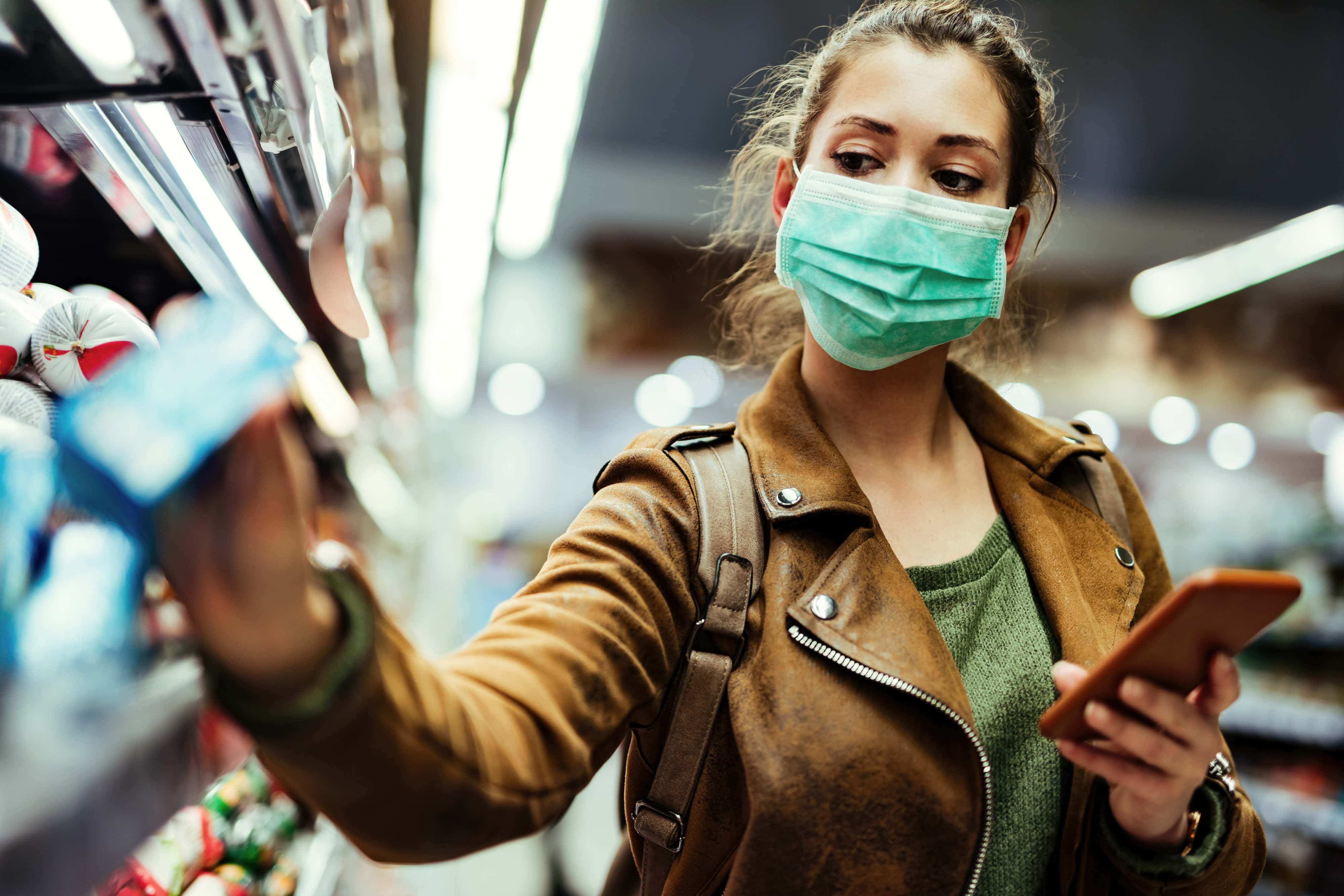 Woman wearing protective mask while using cell phone and buying food in grocery store during virus epidemic.