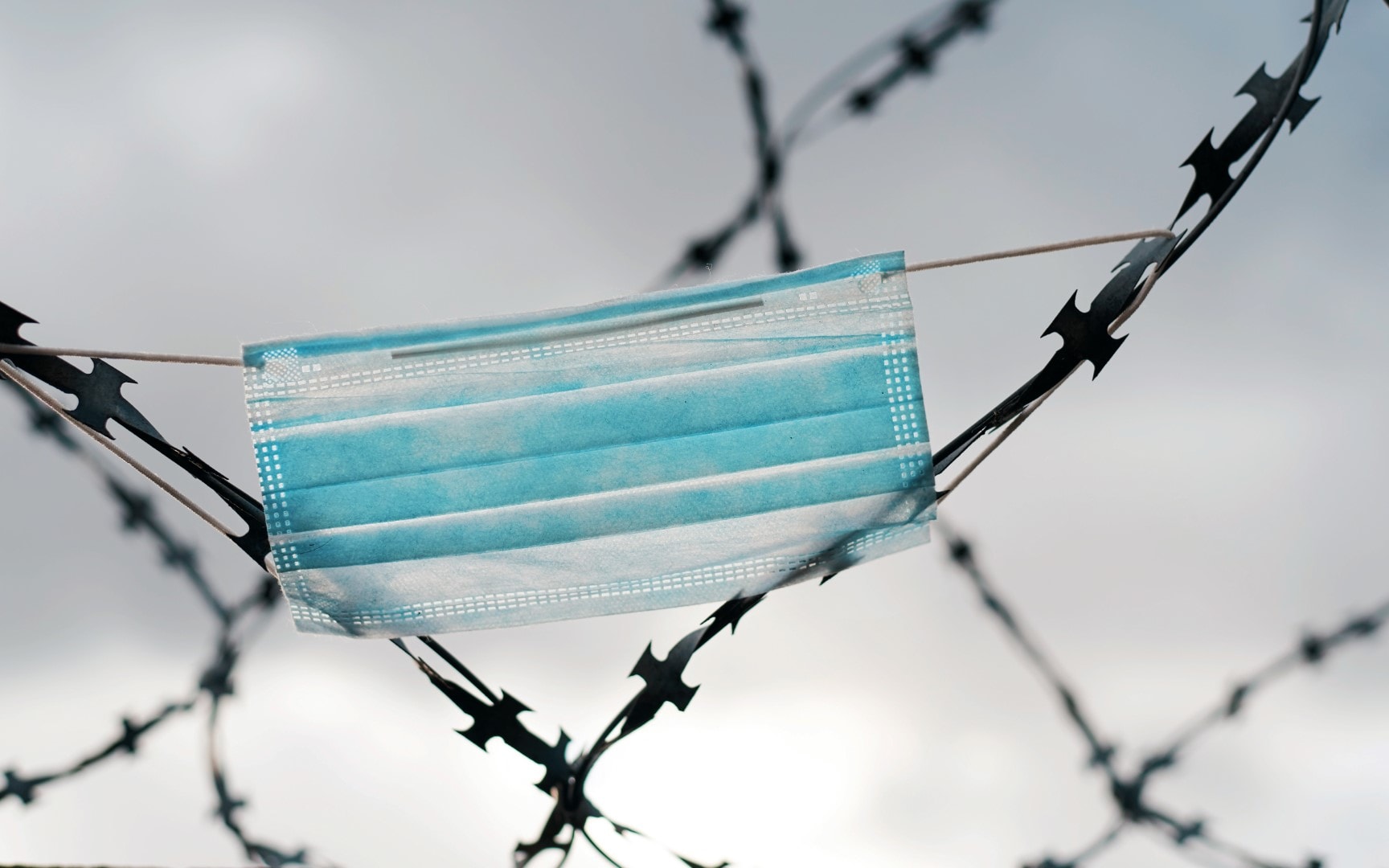 Surgery mask hang on barbed wire, pandemic, isolation and quarantine symbol. Iron fence and personal protective equipment on it.