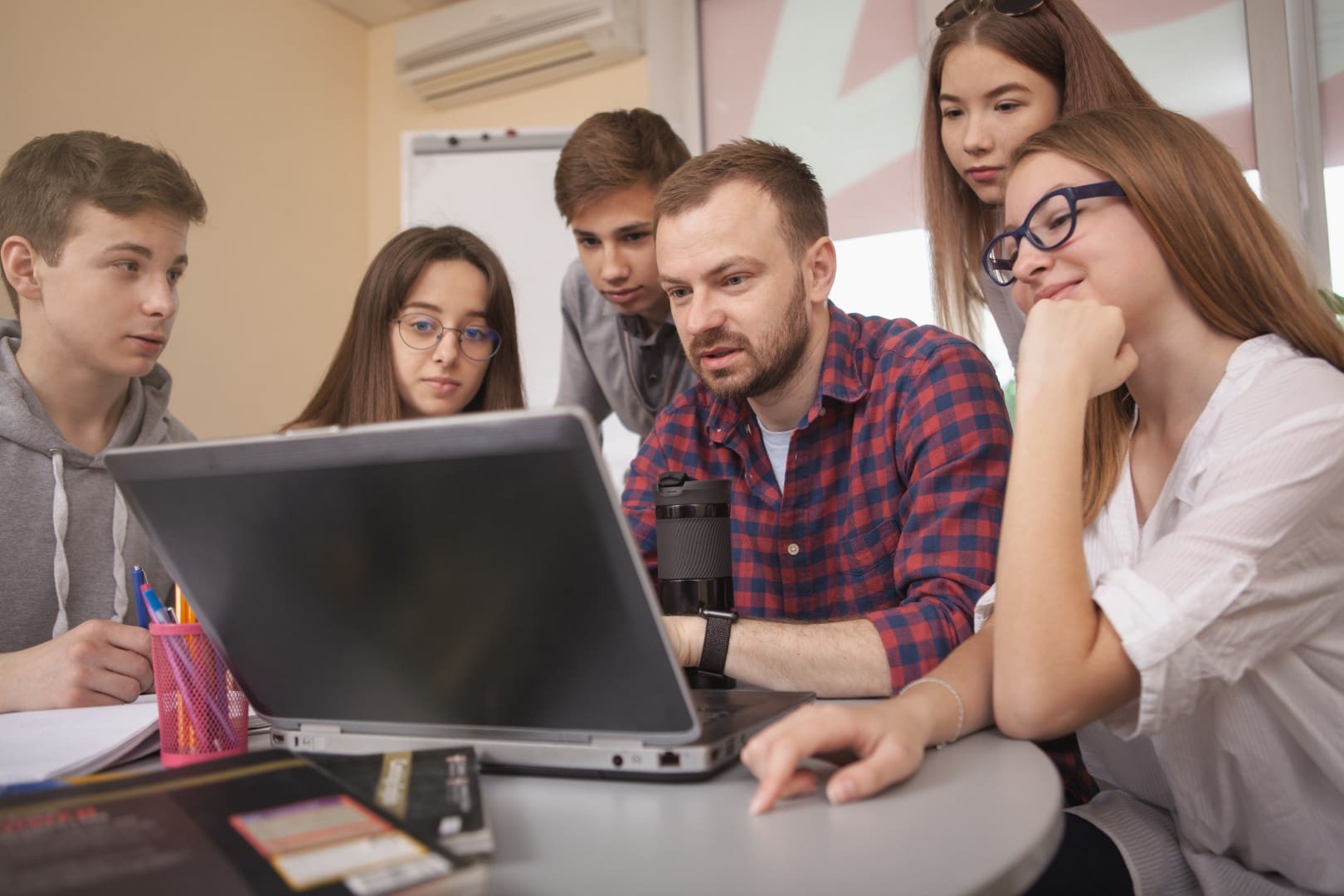 Helpful male teacher using laptop at class, working on a project with his teenage students. Experienced mature mentor working with teenagers at school