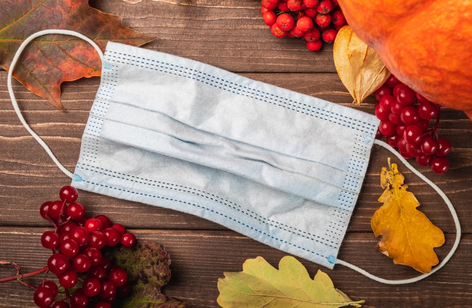 Medical protective mask on an autumn background. Pumpkin, red berries, on a wooden background with a mask in the middle.