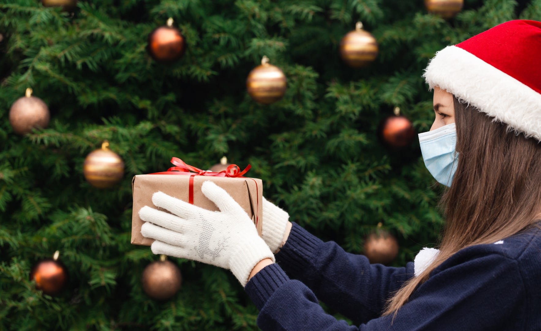 Christmas mask congratulations. Portrait woman wearing santa hat and blue sweater in medical mask, giving gift present box with red ribbon, christmas tree bokeh on background