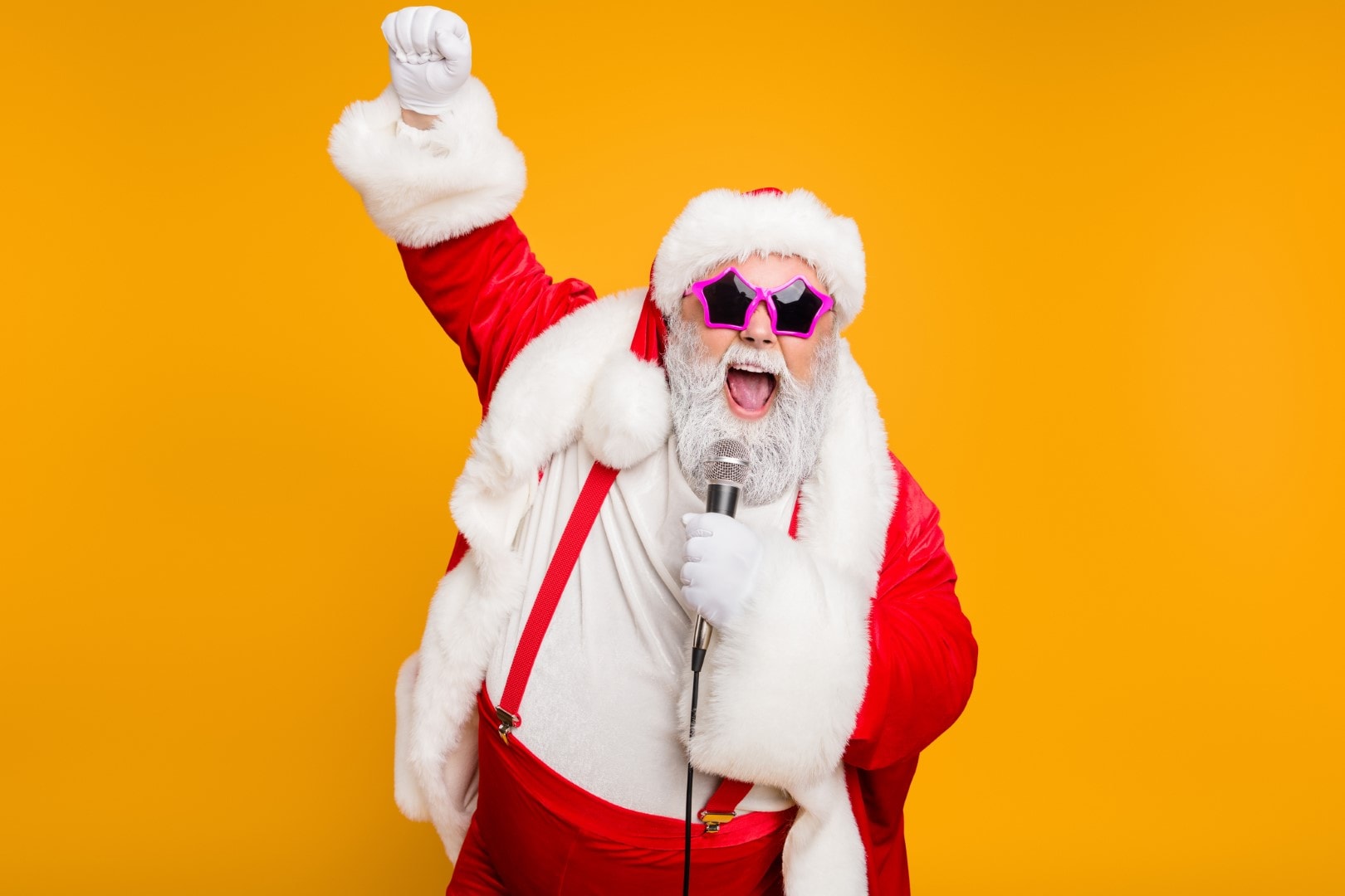 Grey haired stylish christmas grandfather in red hat cap celebrate x-mas party hold microphone sing noel carols feel funky with big belly wear suspenders isolated over yellow color background