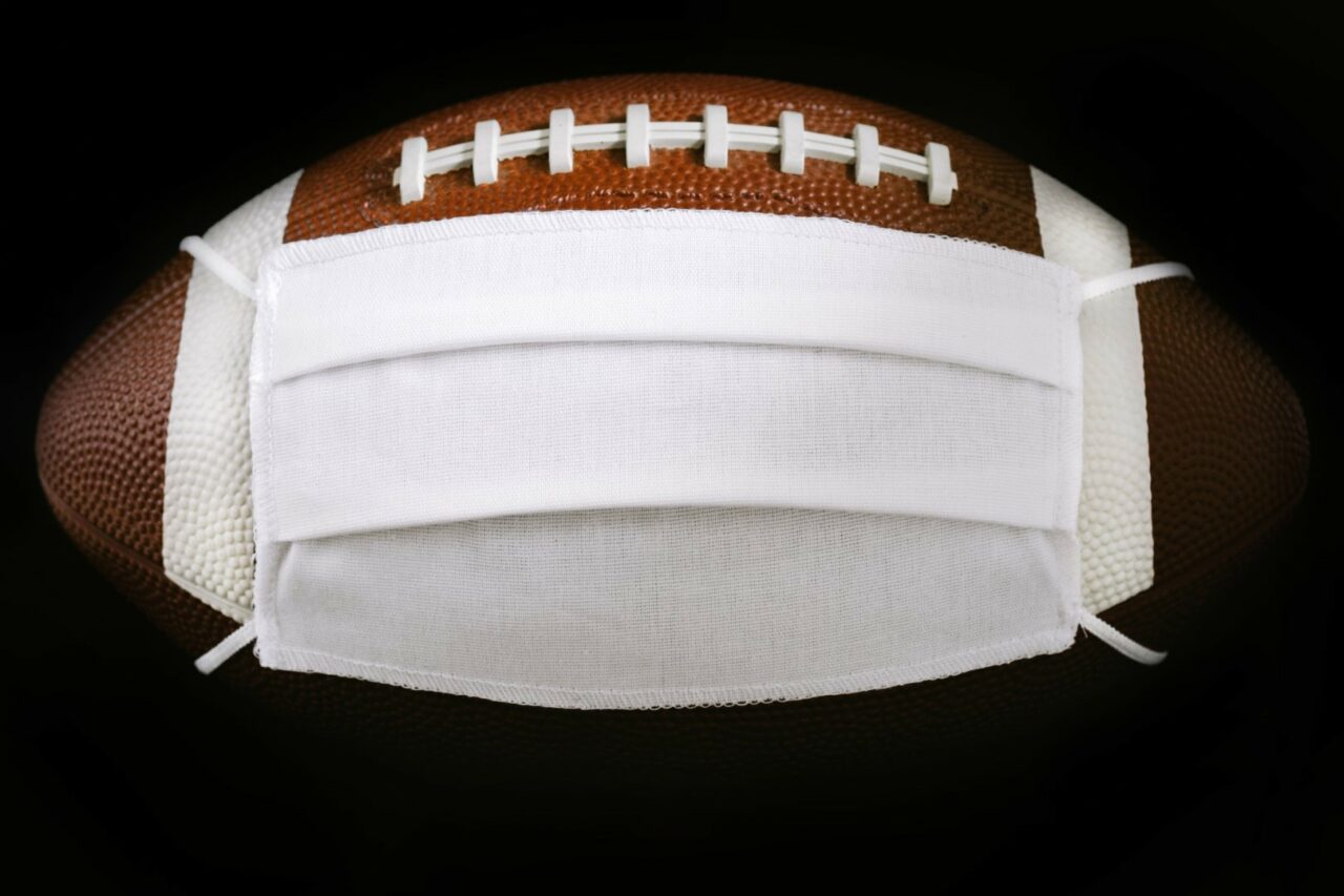 American Football ball in white medical mask for protection of the COVID-19 virus. Cancellation of sporting events and matches concept. Super bowl