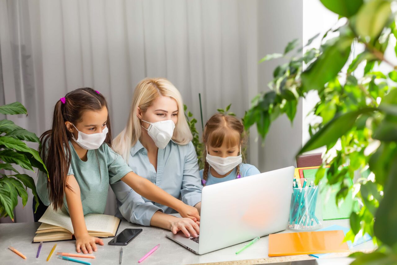Stay home Pandemic coronavirus. Young mother with medical mask trying to freelance remotely on laptop. daughter doing her homework. Remote learning in self-isolation. closed school. Training at home.
