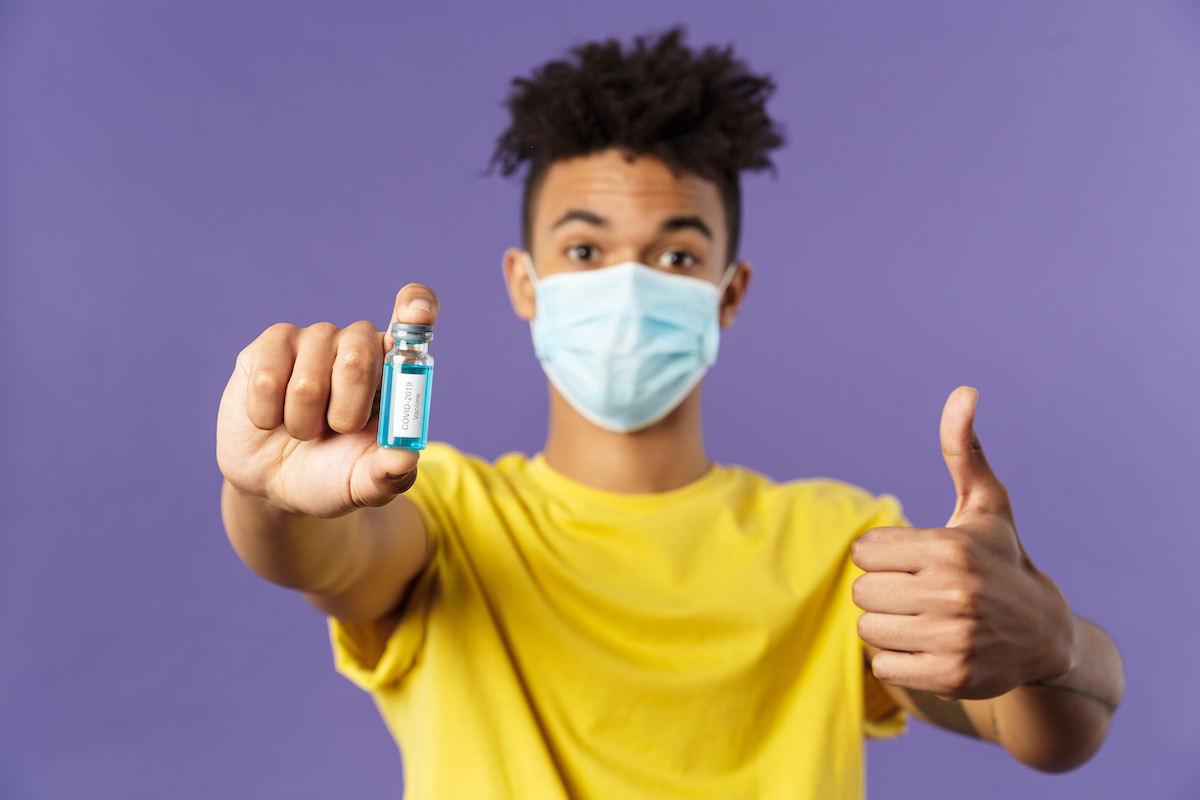 Close-up portrait of upbeat young hispanic man in medical mask holding ampoule with covid19 vaccine, coronavirus drug, show thumbs-up, getting shot at ambulance, standing purple background