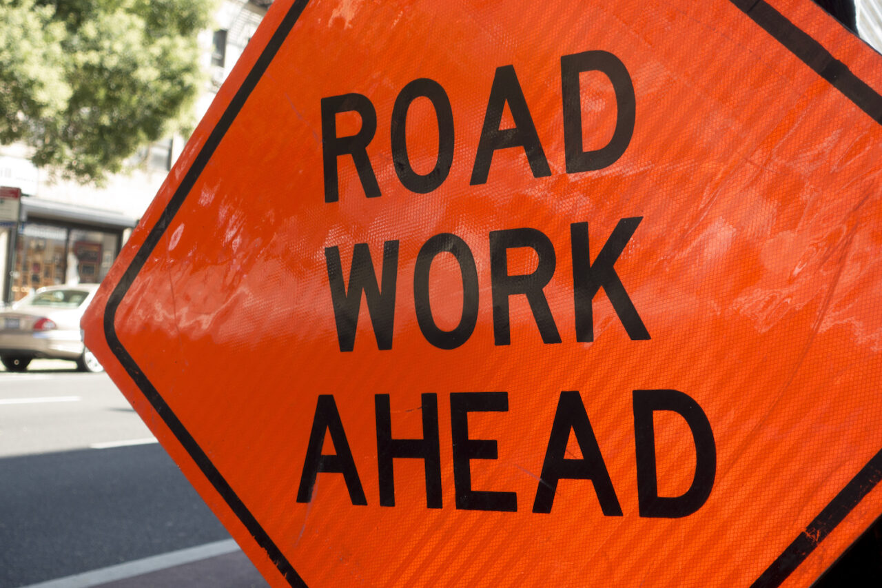 diamond shaped orange red reflector street sign that reads road work ahead