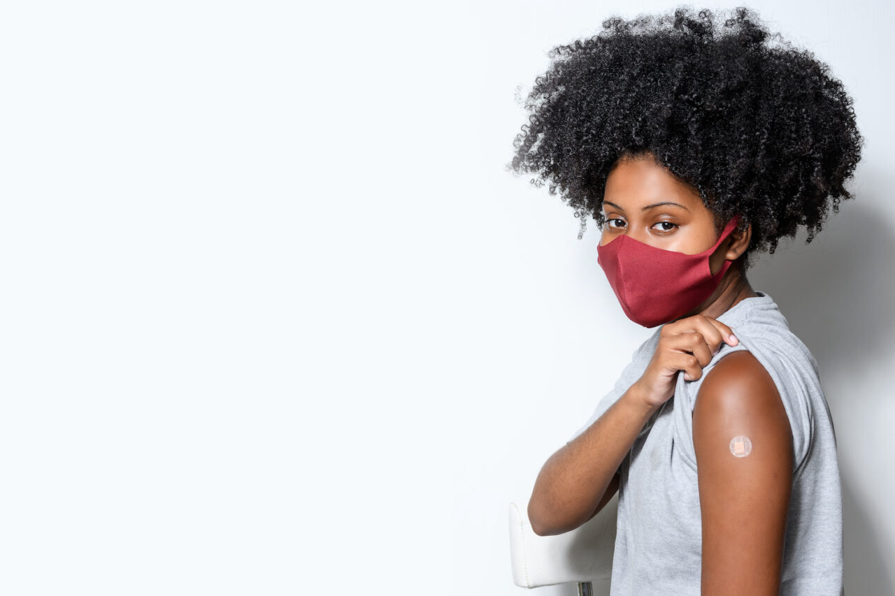black teenager wearing protective mask against covid-19 with a smile on his face shows the vaccine brand, isolated on white background