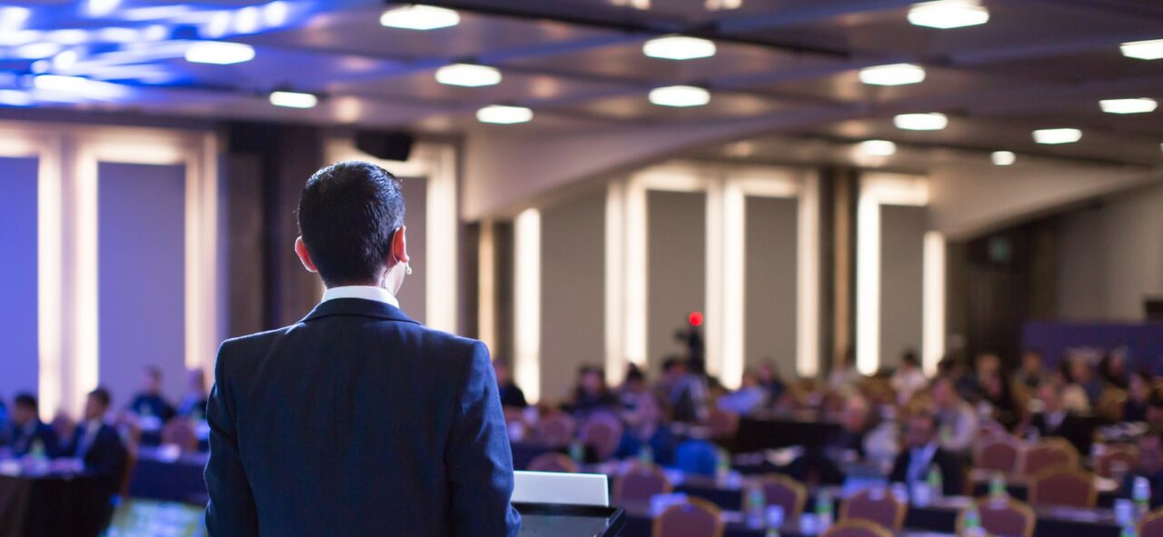 Speaker giving a talk at a corporate business conference. Audience in hall with presenter in front of presentation screen. Corporate executive giving speech during business and entrepreneur seminar.