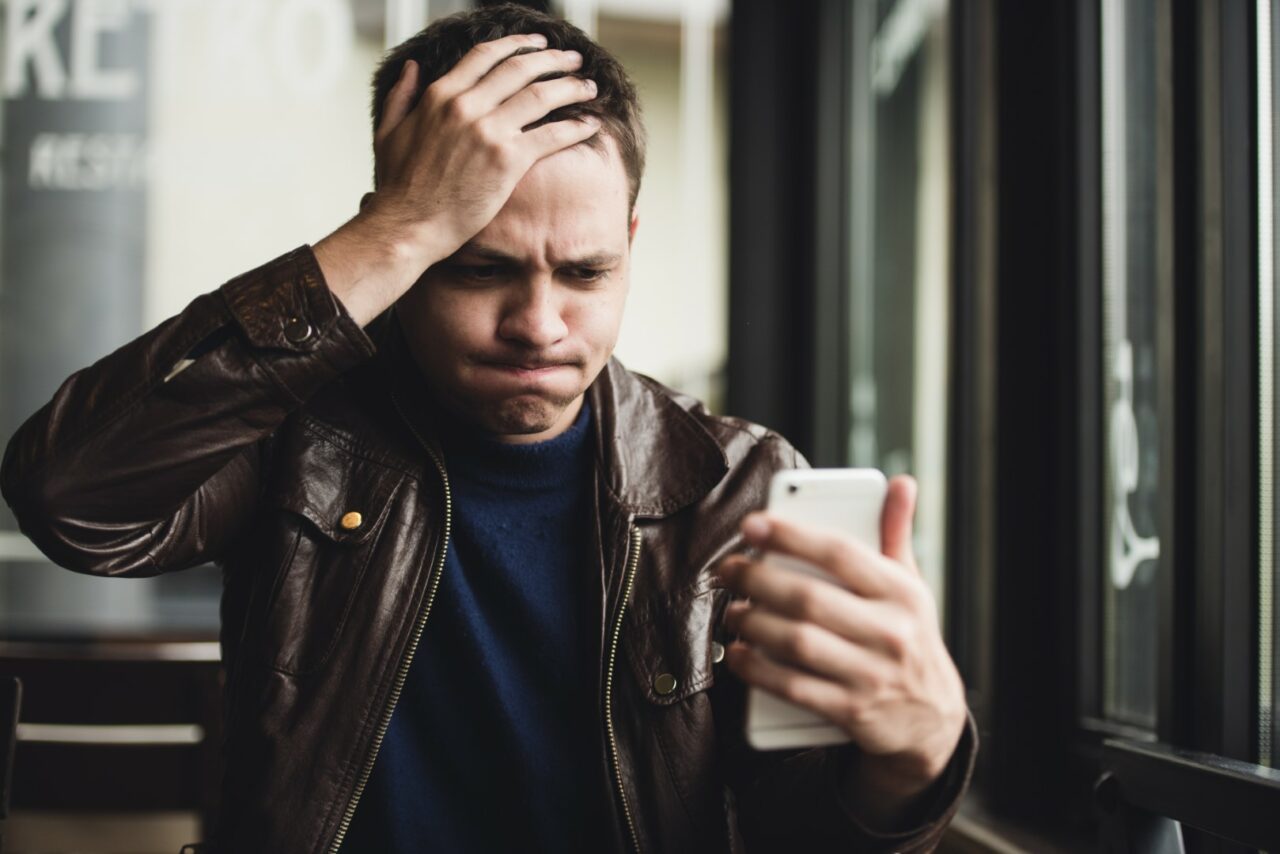 Closeup portrait, stressed young man in purple sweater, shocked surprised, horrified and disturbed, by what he sees on his cell phone, isolated indoors background.