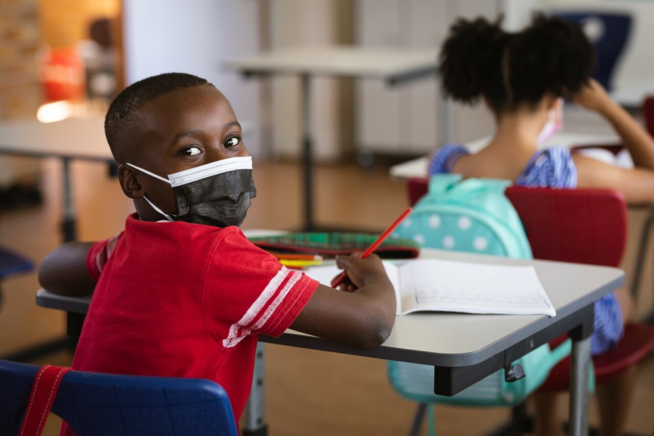 Portrait of african american boy wearing face mask sitting on his desk in class at elementary school