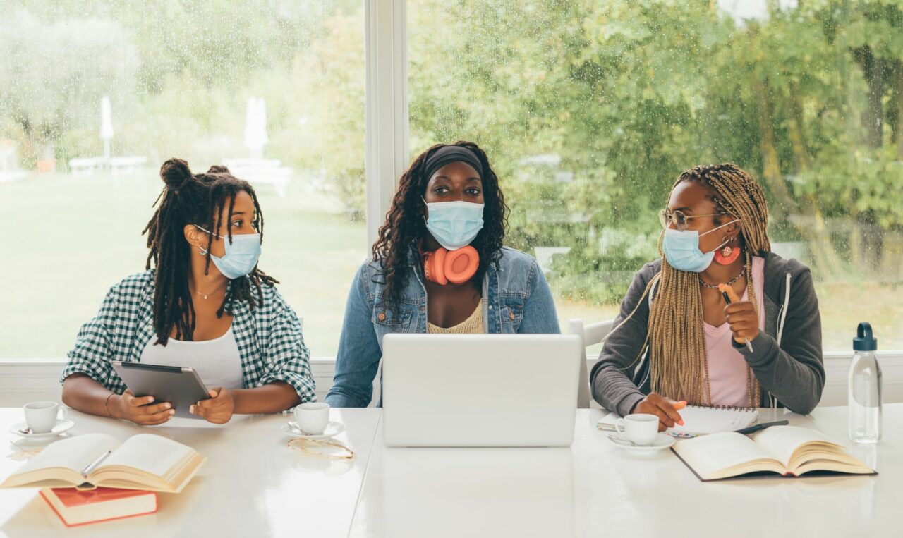 Three African American girls students sitting at the table preparing for exam or making homework together, they are using lap top and digital tablet wearing surgical masks. Back to school concept.