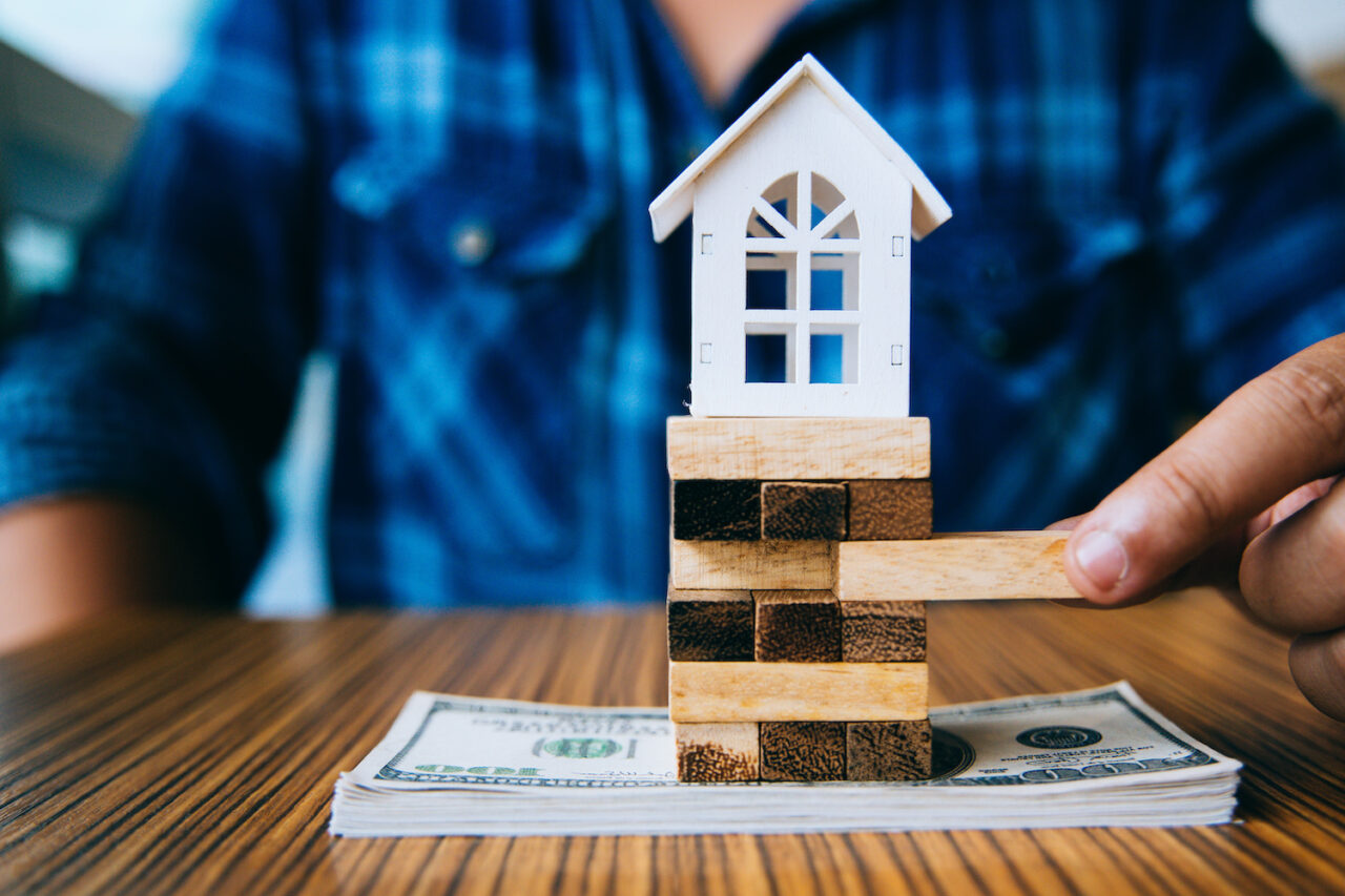 Hand holding a piece of wood block with model white house on dollar banknote. Insurance and property investment real estate concept.