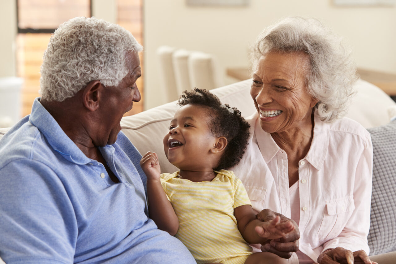 Grandparents Sitting On Sofa At Home Playing With Baby Granddaughter