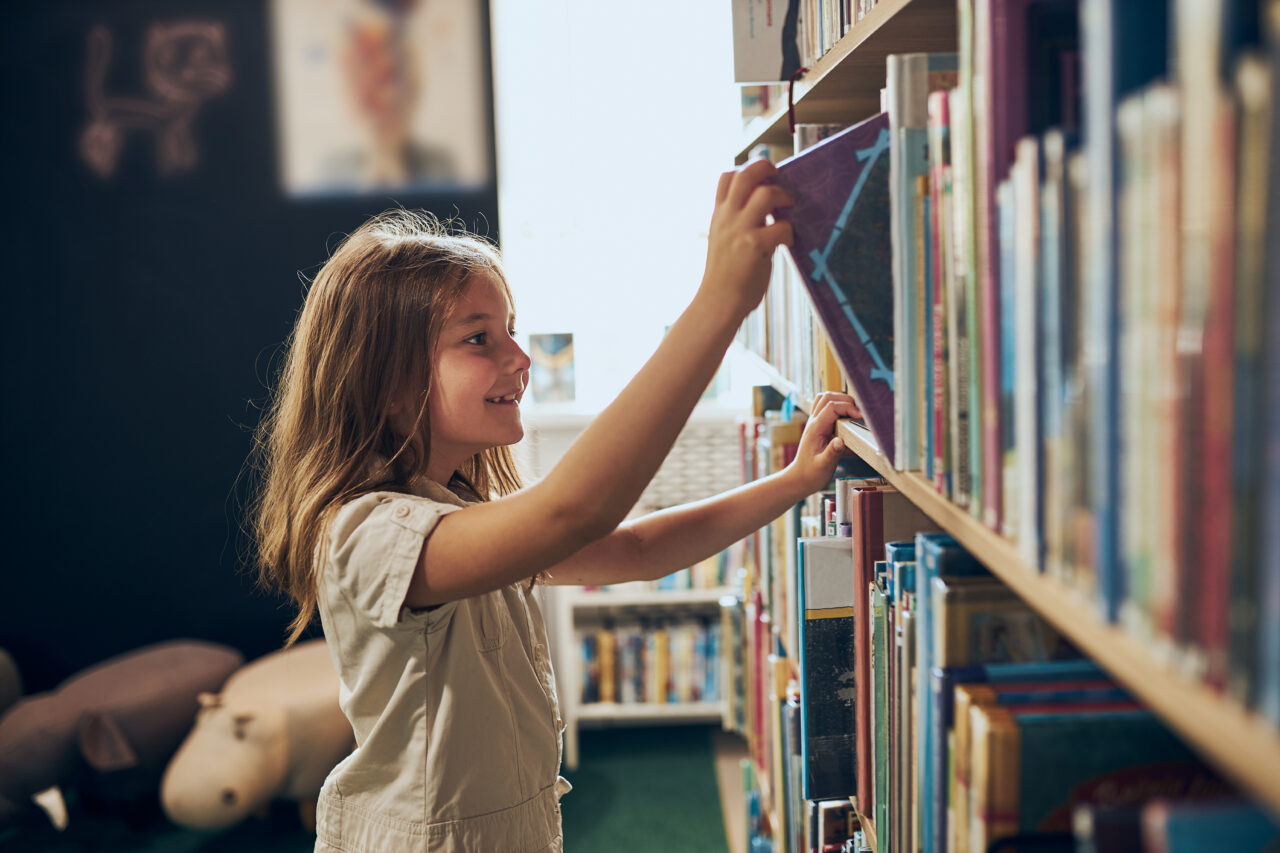 Schoolgirl choosing book in school library. Smart girl selecting literature for reading. Books on shelves in bookstore. Learning from books. School education. Benefits of everyday reading. Child curiosity. Back to school