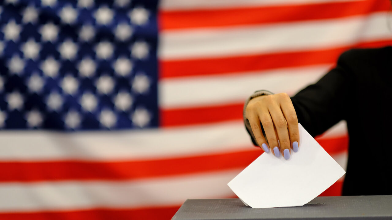 woman putting a ballot in a ballot box on election day. Close up of hand with white votes paper on usa flag background.
