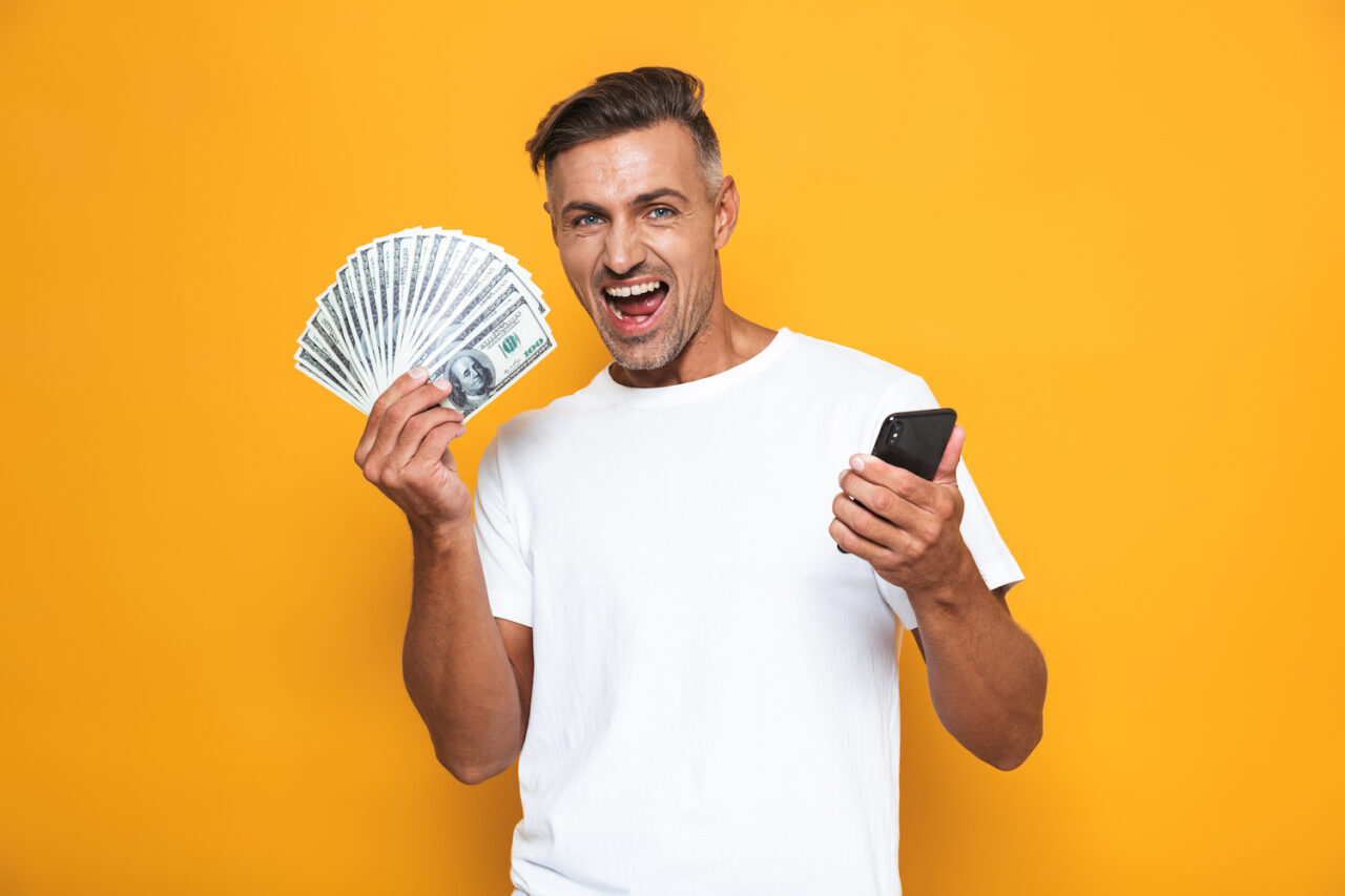 Image of unshaved guy 30s in white t-shirt holding cell phone and bunch of money