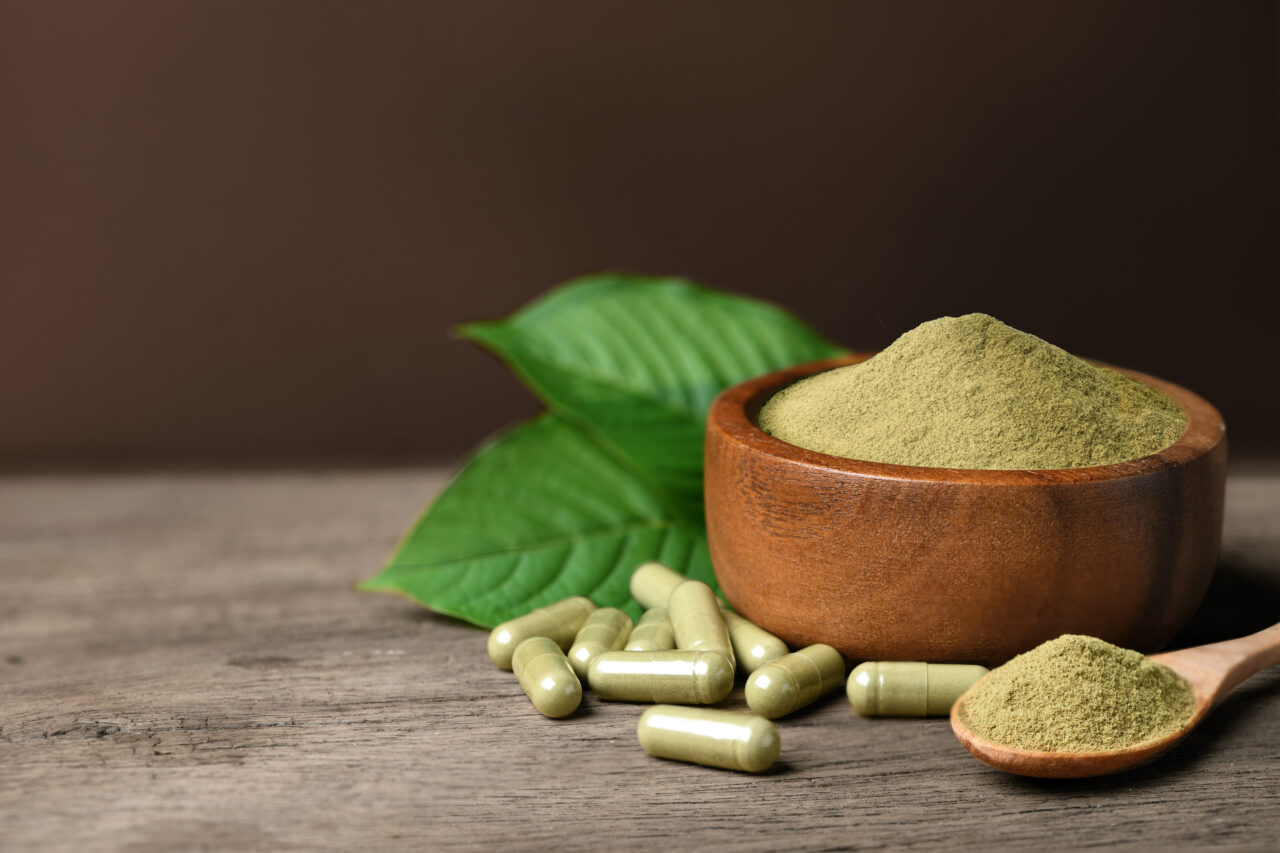 Kratom (Mitragyna speciosa) capsules with powder and fresh leaf on wood background.