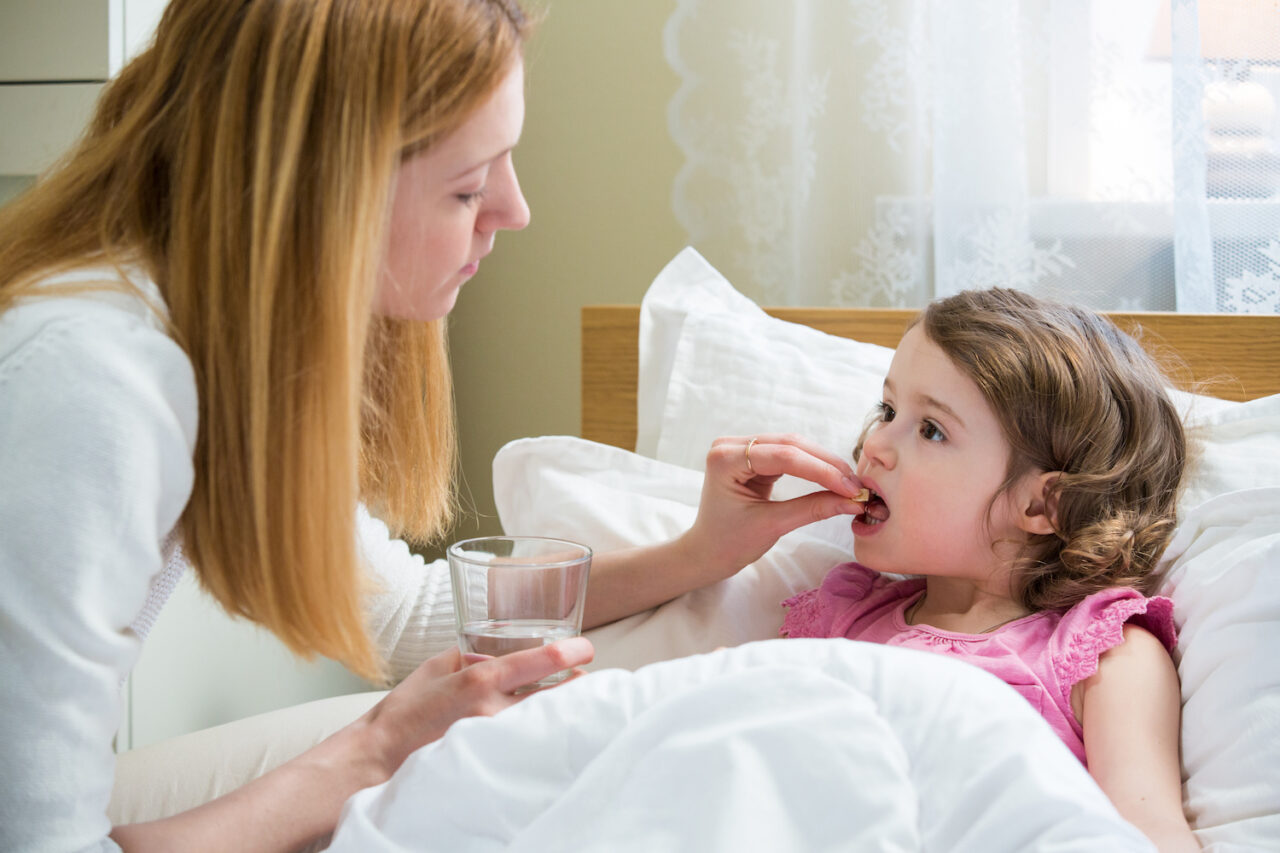 Worried mother giving glass of water to her ill kid. Sick child with high fever laying in bed and taking a medicine. Hand on forehead. Home health care children.