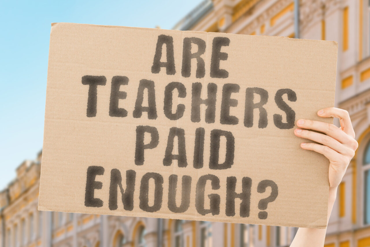 The question " Are teachers paid enough? " is on a banner in men's hands with blurred background. Wealth. Salary. Bank. Banking. Budget. Career. Coin. Corporate. Course. Dollar. Teach. Capital